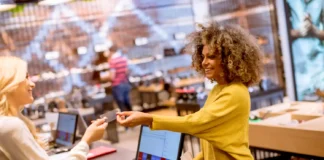 Duas mulheres sorrindo em uma loja ou ponto de venda, trocando um cartão ou cupom promocional, ilustrando ações promocionais criativas para atrair clientes.